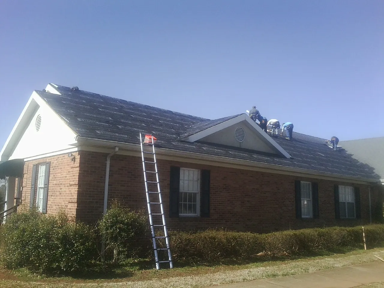 Crew tearing shingles off a brick ranch roof with a ladder set — Emergency Roof Repair work by our crew in What Cheer