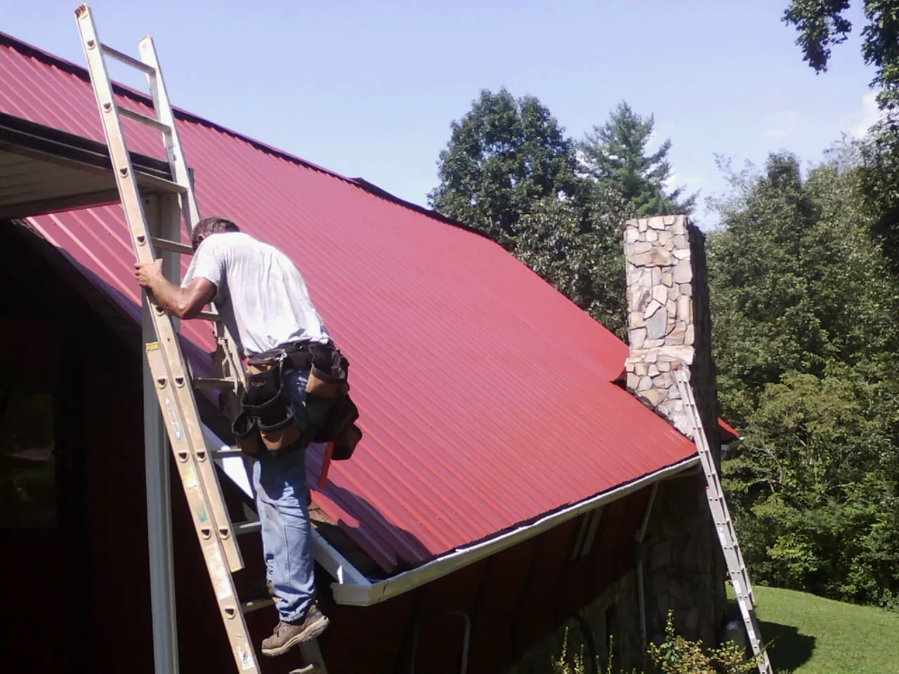 Roofer on a ladder beside a red metal roof and stone chimney — Soffit & Fascia Repair by Roofing Brothers in What Cheer